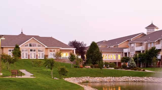 A scenic view of Aberdeen Village senior living facility at dusk, featuring well-maintained green lawns, a small pond with a rocky edge, several trees, a walking path with a bench, and beige multi-story buildings with large windows and balconies.