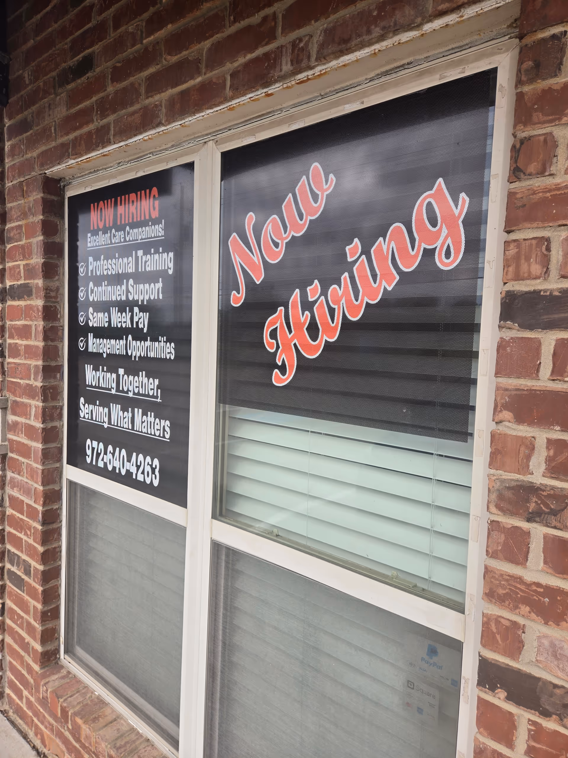 Brick building exterior with a window displaying a Now Hiring sign.