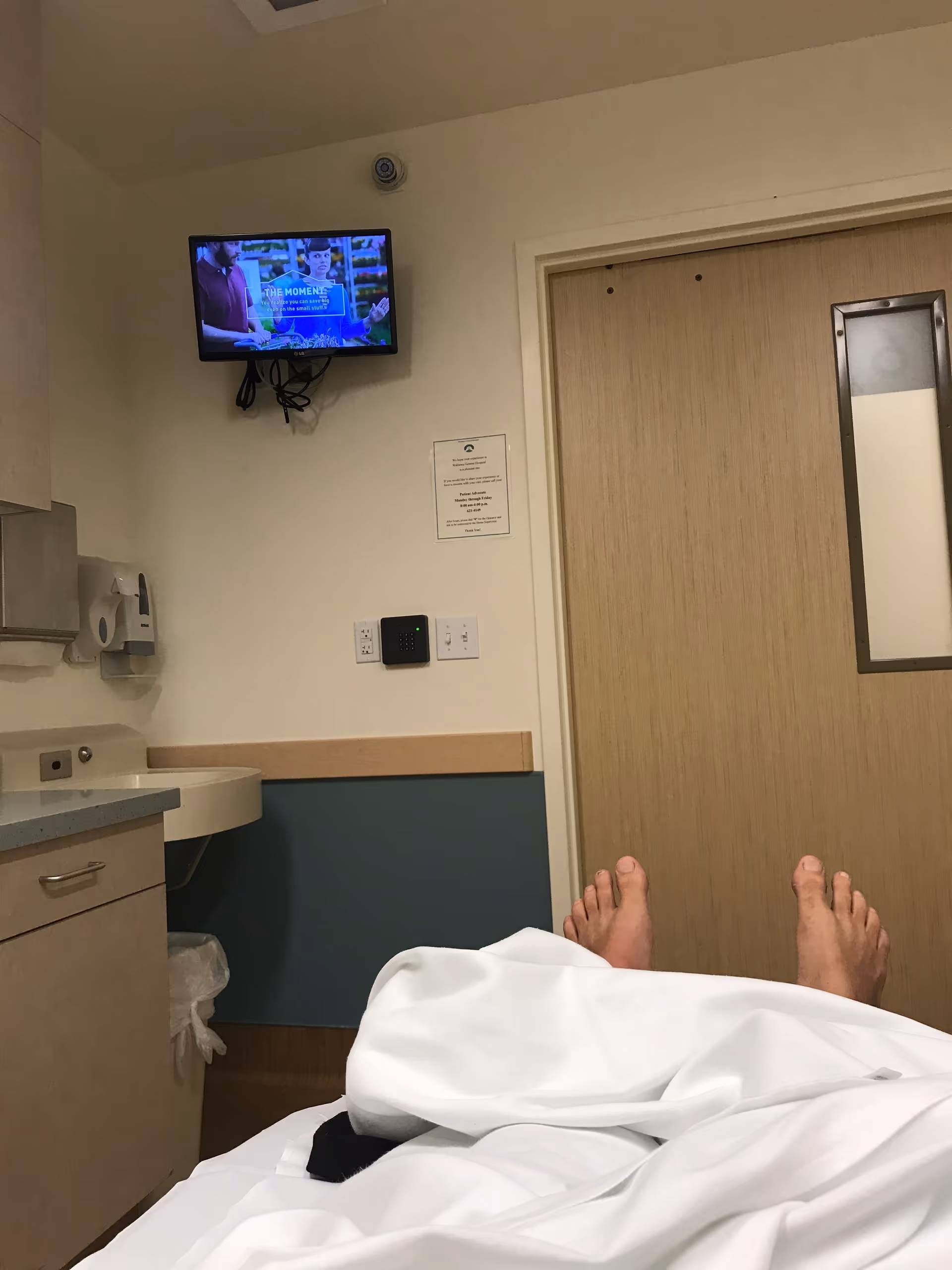 View from a patient bed with a wall-mounted TV, sink, and wooden door.