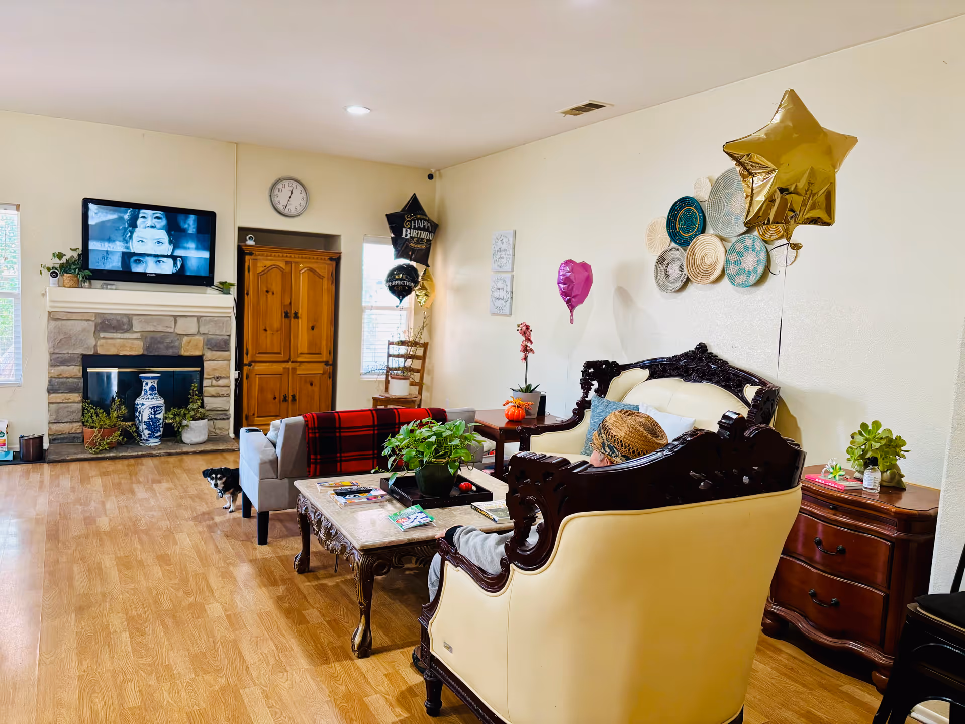 Living room in a senior facility featuring ornate furniture, balloons, TV, fireplace, and potted plants.