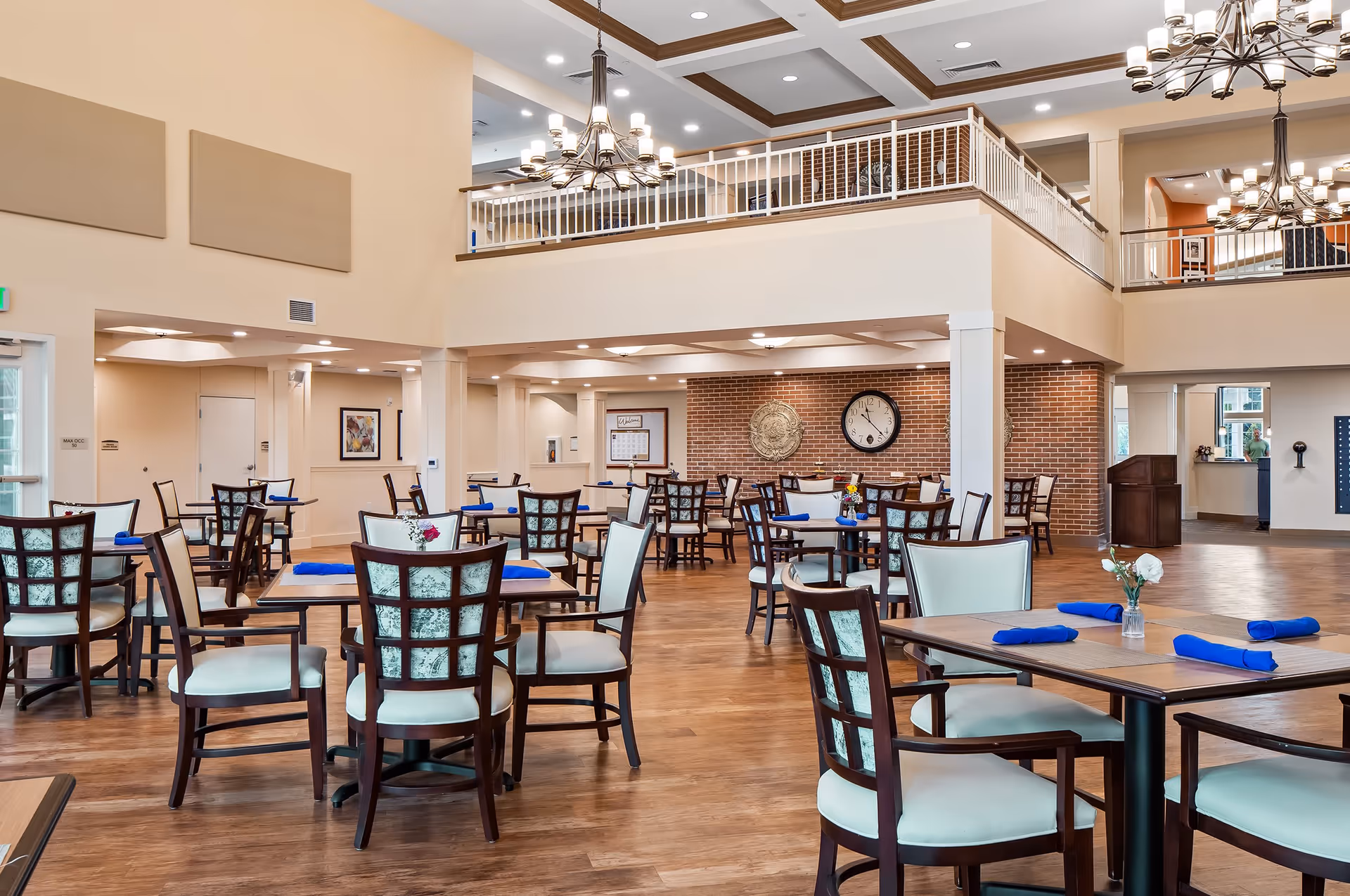 Spacious dining room with multiple wooden tables and chairs with light-colored cushions. Each table is set with blue napkins and small flower vases. The room features high ceilings with chandeliers, a second-floor balcony with white railings, and a brick accent wall with a large clock and decorative medallions.