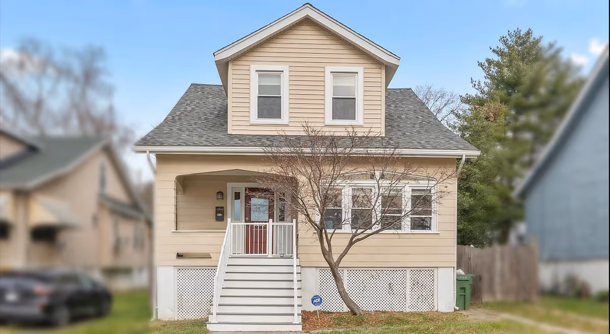 Beige two-story house with front porch, stairs, and leafless tree in front yard.
