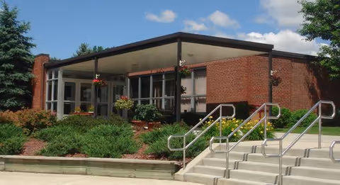 Brick Antwerp Manor assisted-living facility exterior with glass entry, stairs, and landscaped frontage.