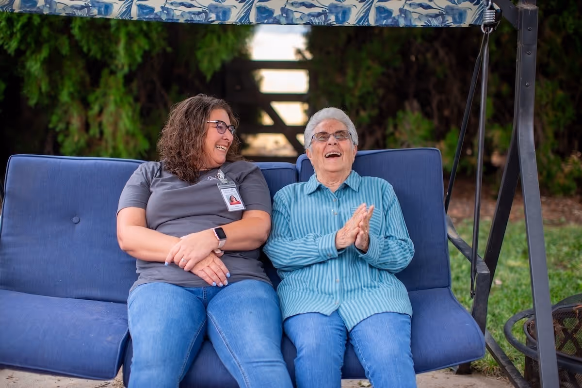 Two women sit on a blue outdoor swinging bench, smiling and laughing together.
