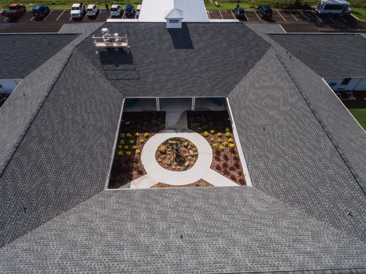 Aerial view of a building with a central courtyard garden featuring a circular walkway and landscaped plants. The building has a gray shingled roof and a small cupola on top. Several cars are parked in the parking lot surrounding the building.