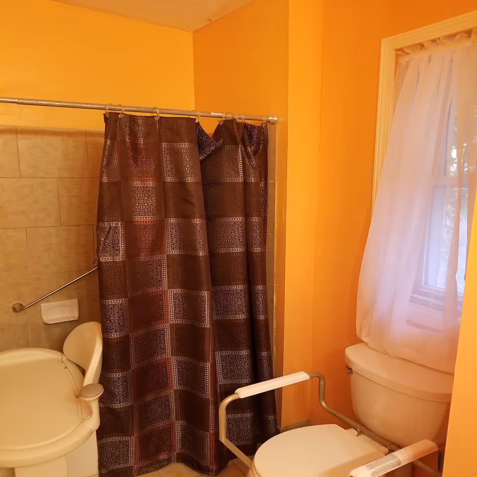 Orange-walled bathroom with shower curtain, toilet, and window in a senior living facility.