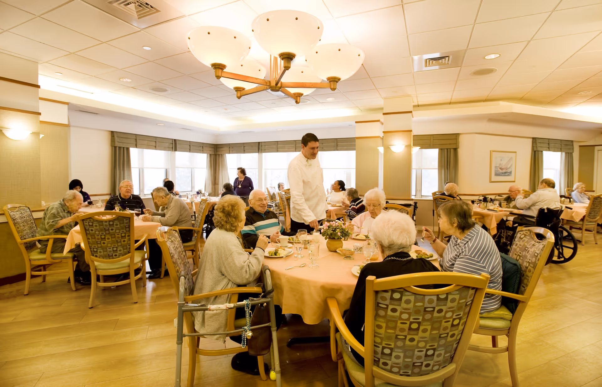 Elderly residents sit at several round tables in a bright communal dining room while a staff member serves them.