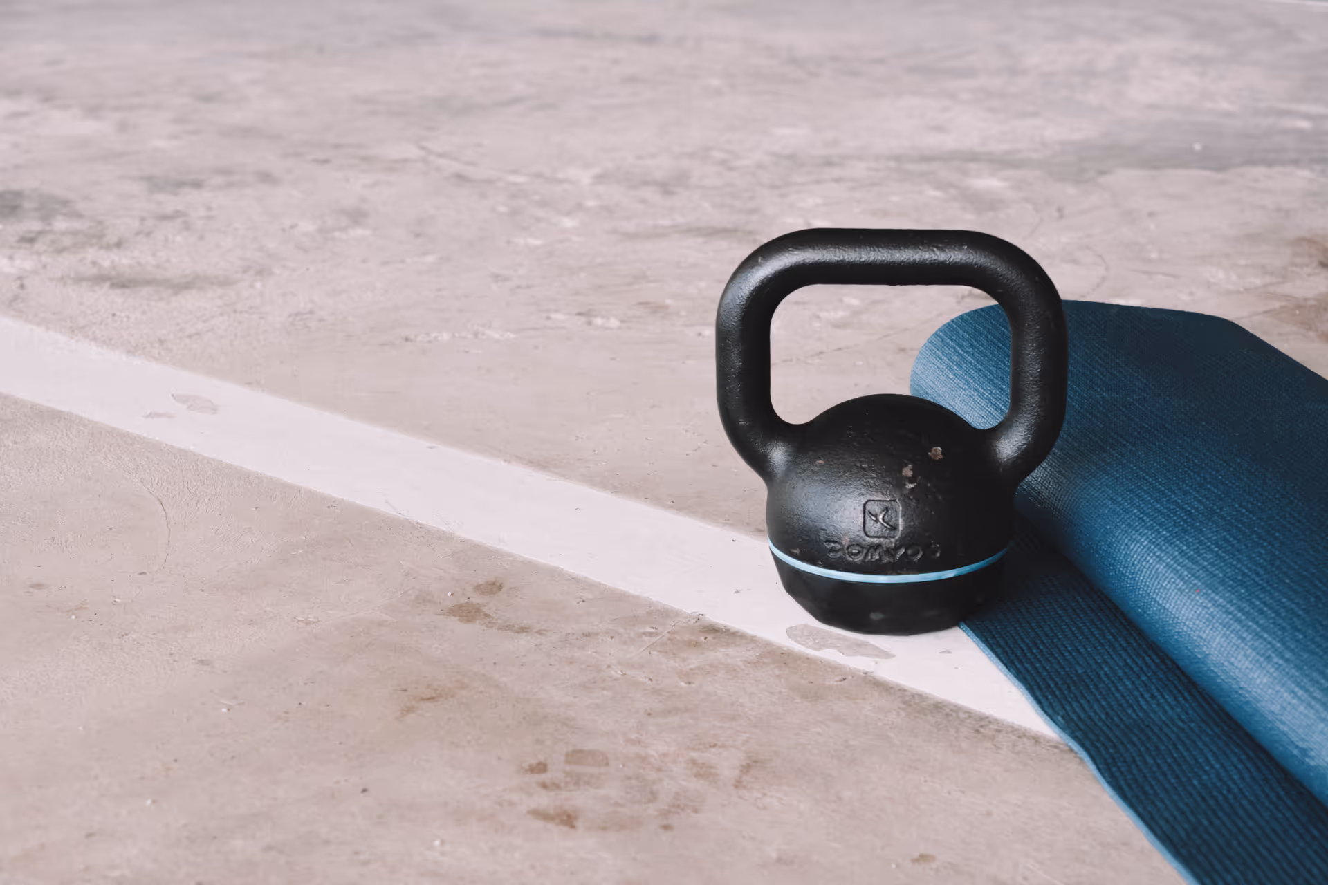 A black kettlebell rests on a blue yoga mat on a concrete floor indoors.