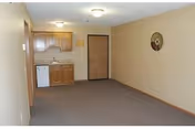 Empty beige living area with a small kitchenette, doorway, and clock on the wall.