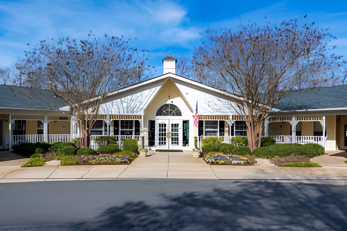 Front entrance of a single-story building with a white gabled entry, porches on each side, landscaped beds and an American flag.