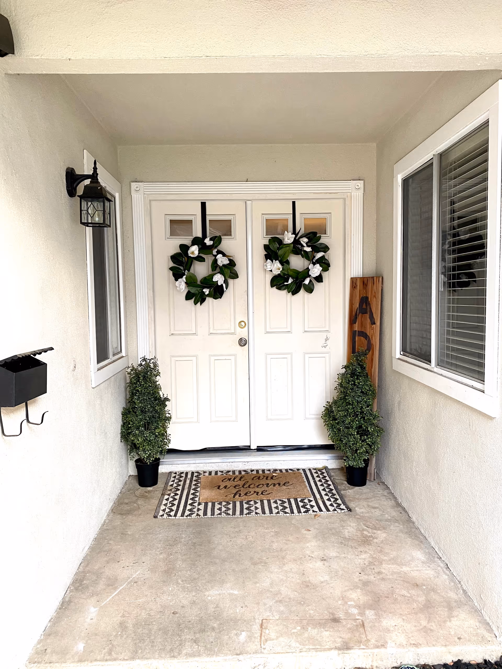 Front entrance with double white doors, decorative wreaths, a welcome mat, and potted plants.