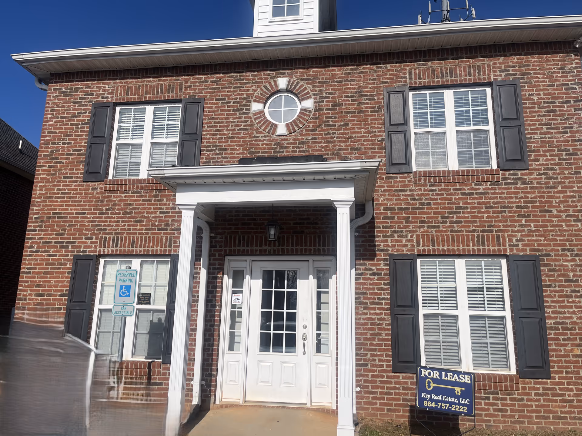 Brick two-story building entrance with white columns, black shutters, and a For Lease sign.