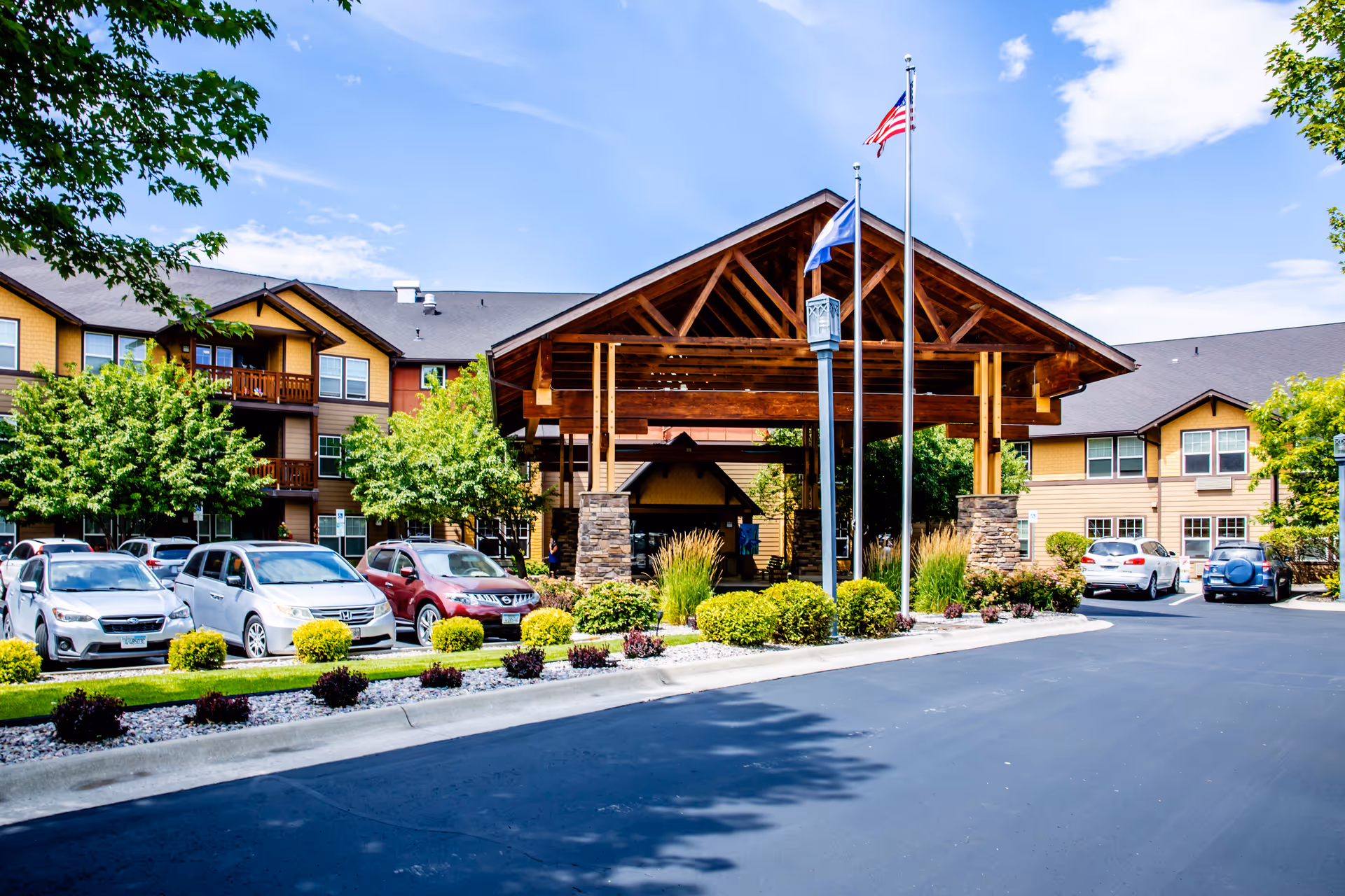 Front entrance of a senior living facility with a large wooden porte-cochere, flagpoles, landscaped beds, and parked cars.