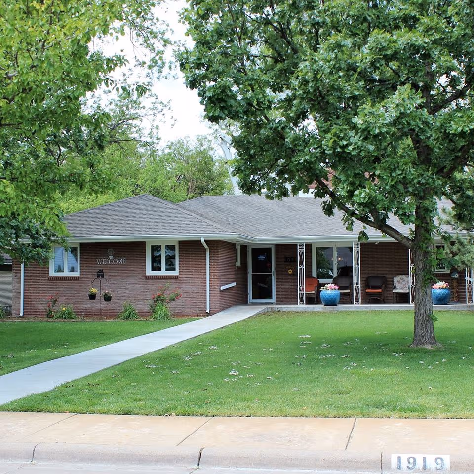 A single-story brick senior living facility with a front porch, lawn, and large trees.
