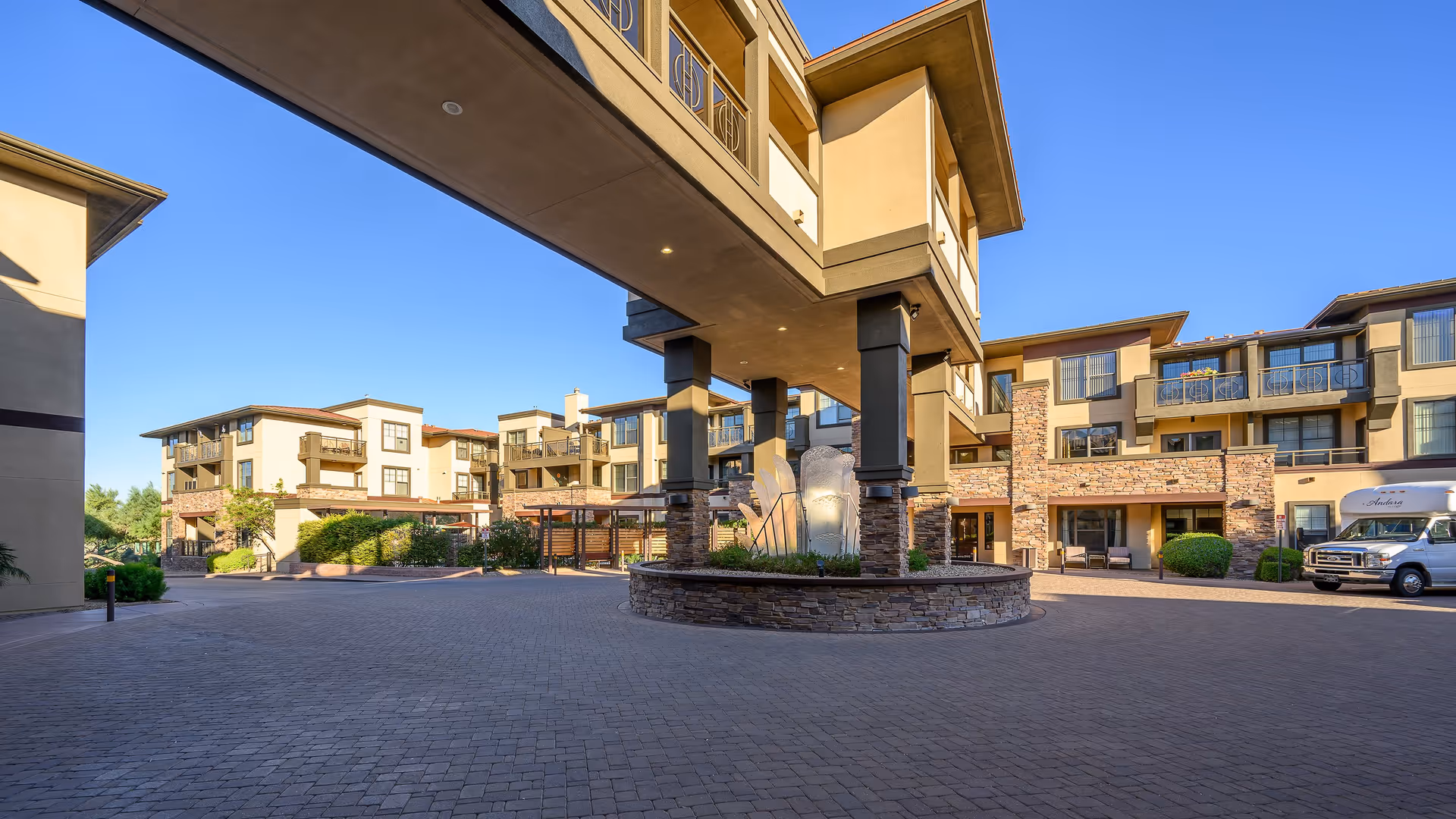 Exterior view of a senior living facility with multiple three-story buildings featuring balconies and stone accents. The image shows a paved courtyard area with a covered entrance supported by columns and a decorative stone planter in the center. A vehicle is parked on the right side near the building.