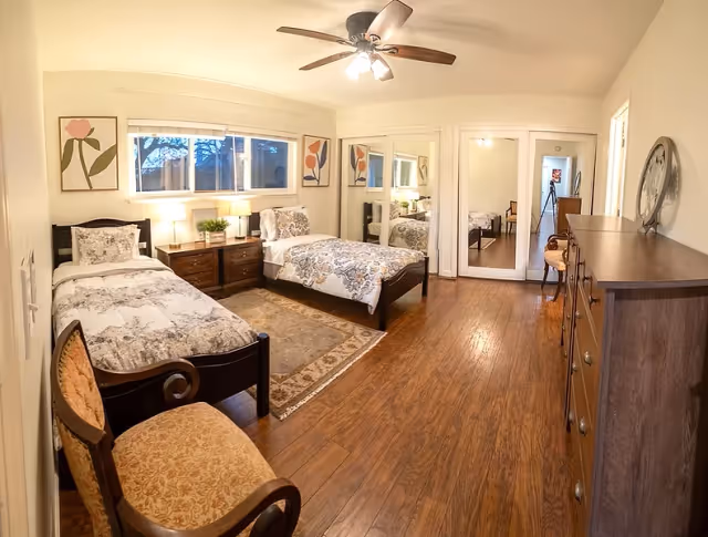Bright bedroom with two twin beds, wooden nightstand and dresser, chairs, mirrored closet doors, ceiling fan, and hardwood floor.