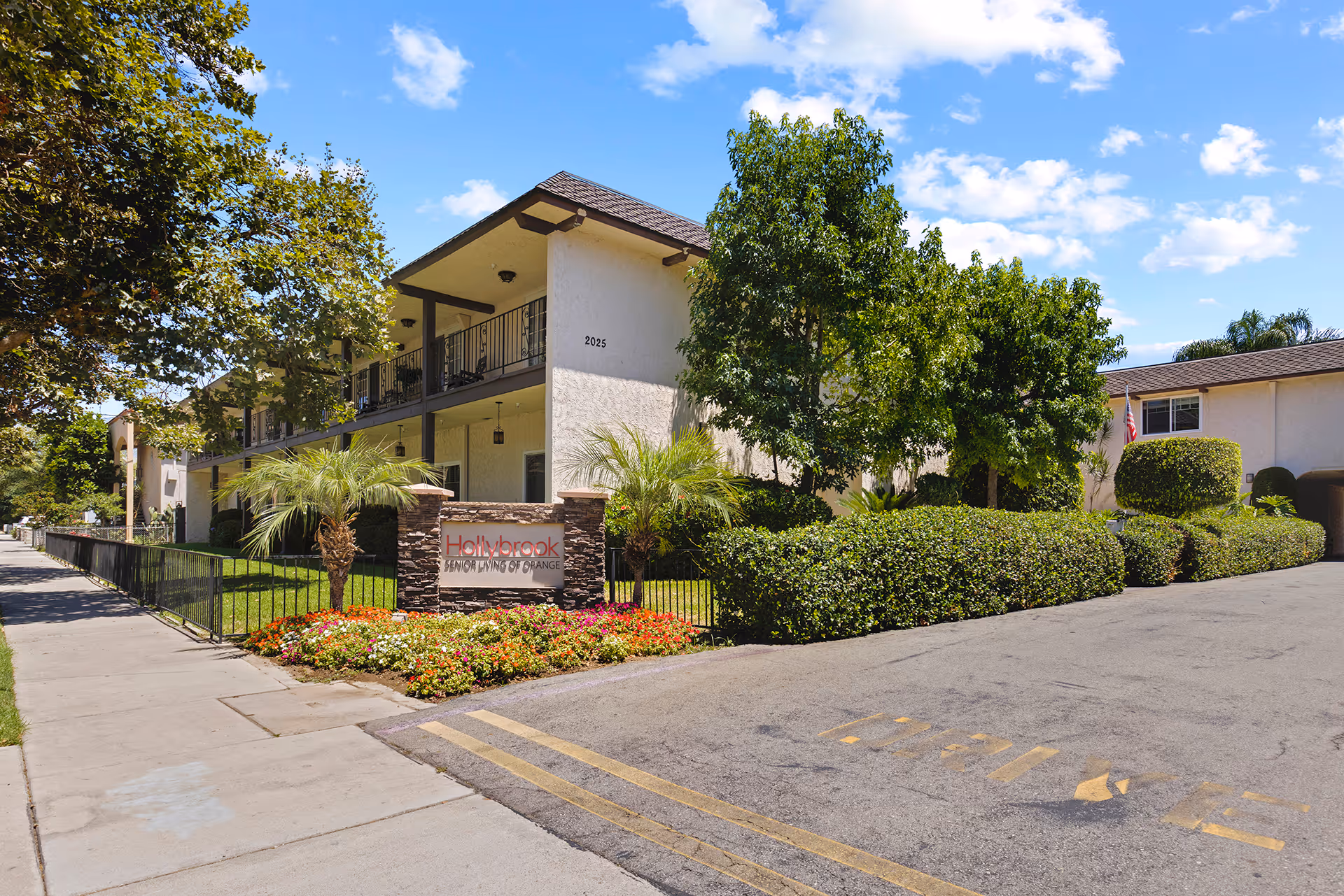 Exterior view of Hollybrook Senior Living of Orange facility showing a two-story building with balconies, surrounded by trees, bushes, and a flower bed. A sign with the facility name is visible near the entrance along a paved driveway and sidewalk under a partly cloudy blue sky.