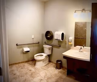 Wheelchair-accessible restroom with a toilet and grab bars, wall-mounted paper dispenser, sink with mirror and tiled floor.