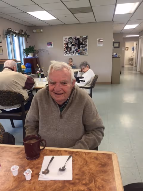 An elderly man smiling at a table with a mug and utensils in a communal dining room, with other seniors seated and a bulletin board on the wall.