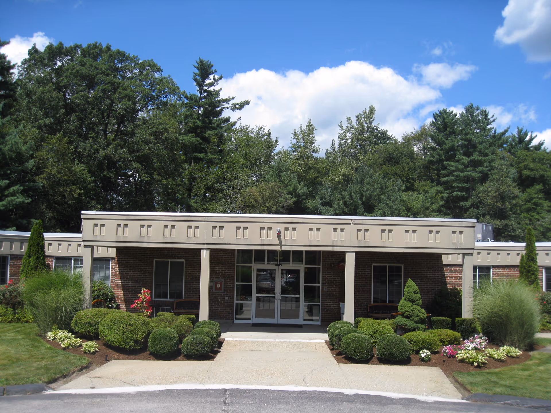 Front exterior view of South Shore Rehabilitation & Skilled Care Center building with a flat roof, brick walls, large glass entrance doors, and well-maintained landscaping including bushes, flowers, and trees under a blue sky with some clouds.
