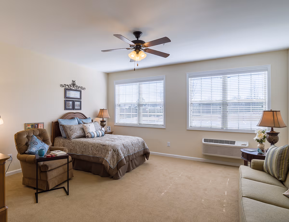 A furnished senior living bedroom with bed, seating, windows, and a ceiling fan.