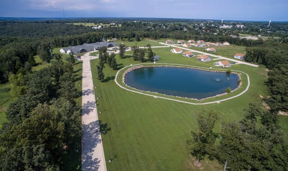 Aerial view of a senior living campus with a circular pond, cottages, and extensive green grounds.
