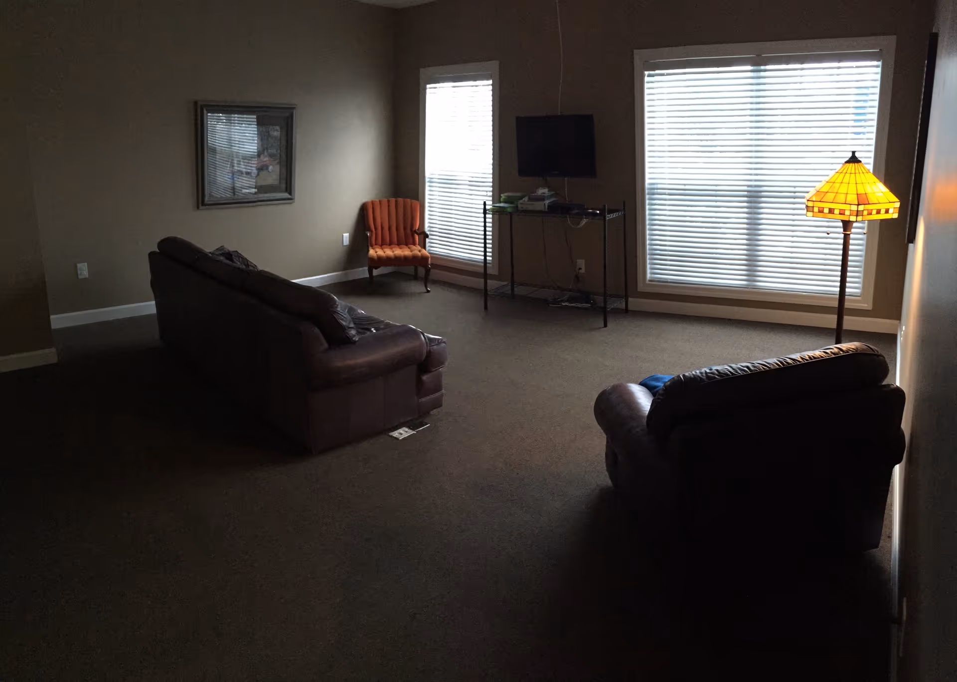 A dimly lit living room with two brown leather sofas facing a small TV on a metal stand. There is an orange upholstered chair in the corner near two large windows with blinds. A floor lamp with a yellow stained-glass shade is on the right side of the room. A framed picture hangs on the wall above the orange chair.