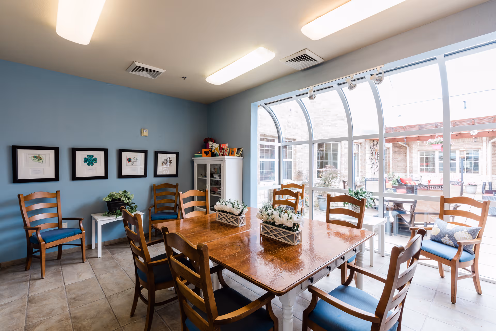 A bright dining room with a large wooden table surrounded by eight wooden chairs with blue cushions. The room has light blue walls adorned with four framed pictures. There is a white cabinet in the corner with decorative items on top. Large windows and a glass ceiling provide a view of an outdoor patio area with additional seating and plants.