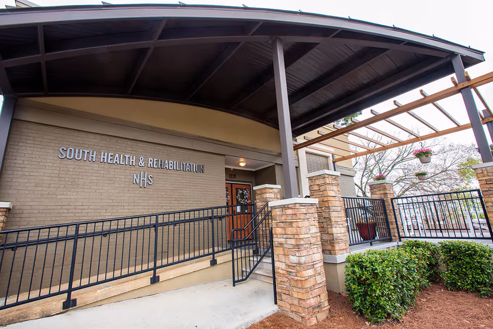 Entrance of South Health & Rehabilitation facility with a covered porch, brick pillars, black metal railings, and a ramp leading to a wooden door. There are hanging flower pots and some greenery around the entrance.