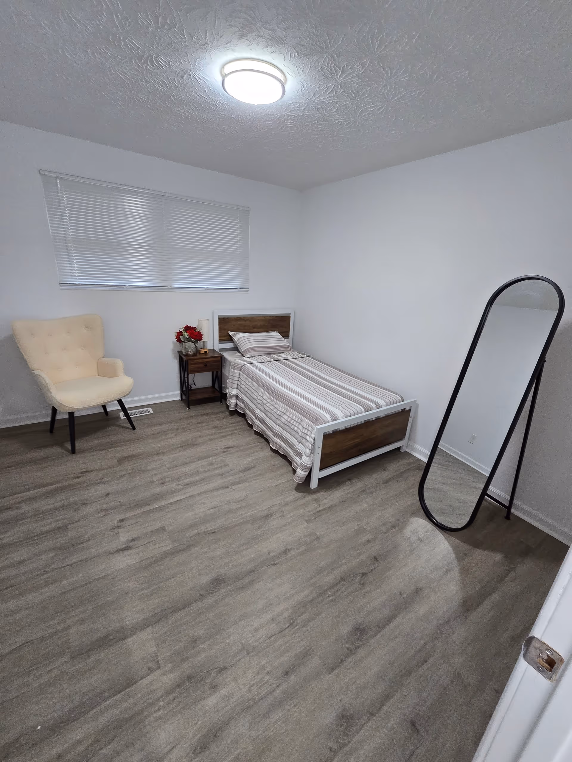 A simple bedroom with single bed, nightstand, chair, and a standing full-length mirror.
