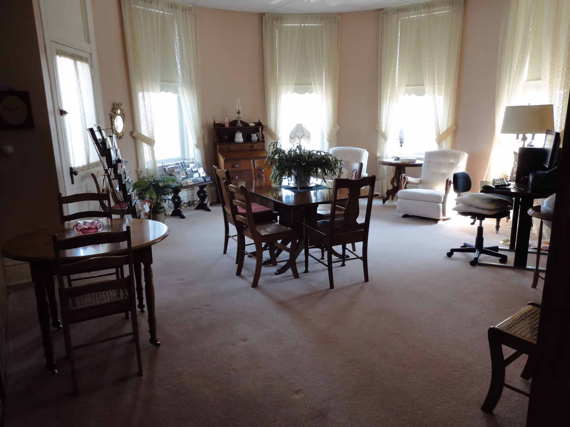 A spacious, well-lit room with multiple wooden tables and chairs, a potted plant on the central table, two white upholstered armchairs near windows with sheer curtains, a desk with a computer and lamp, and a wooden cabinet with decorative items.