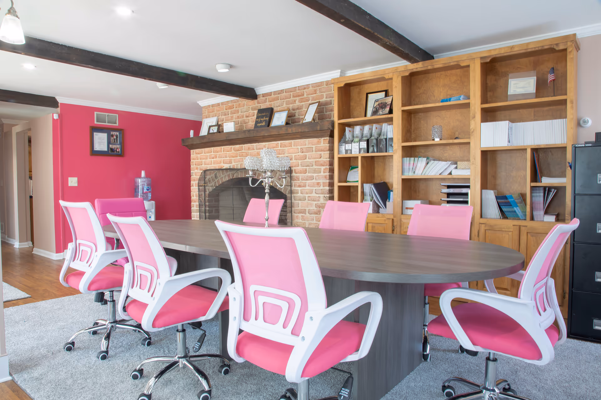 Bright dining room with pink chairs around a large oval table and brick fireplace.