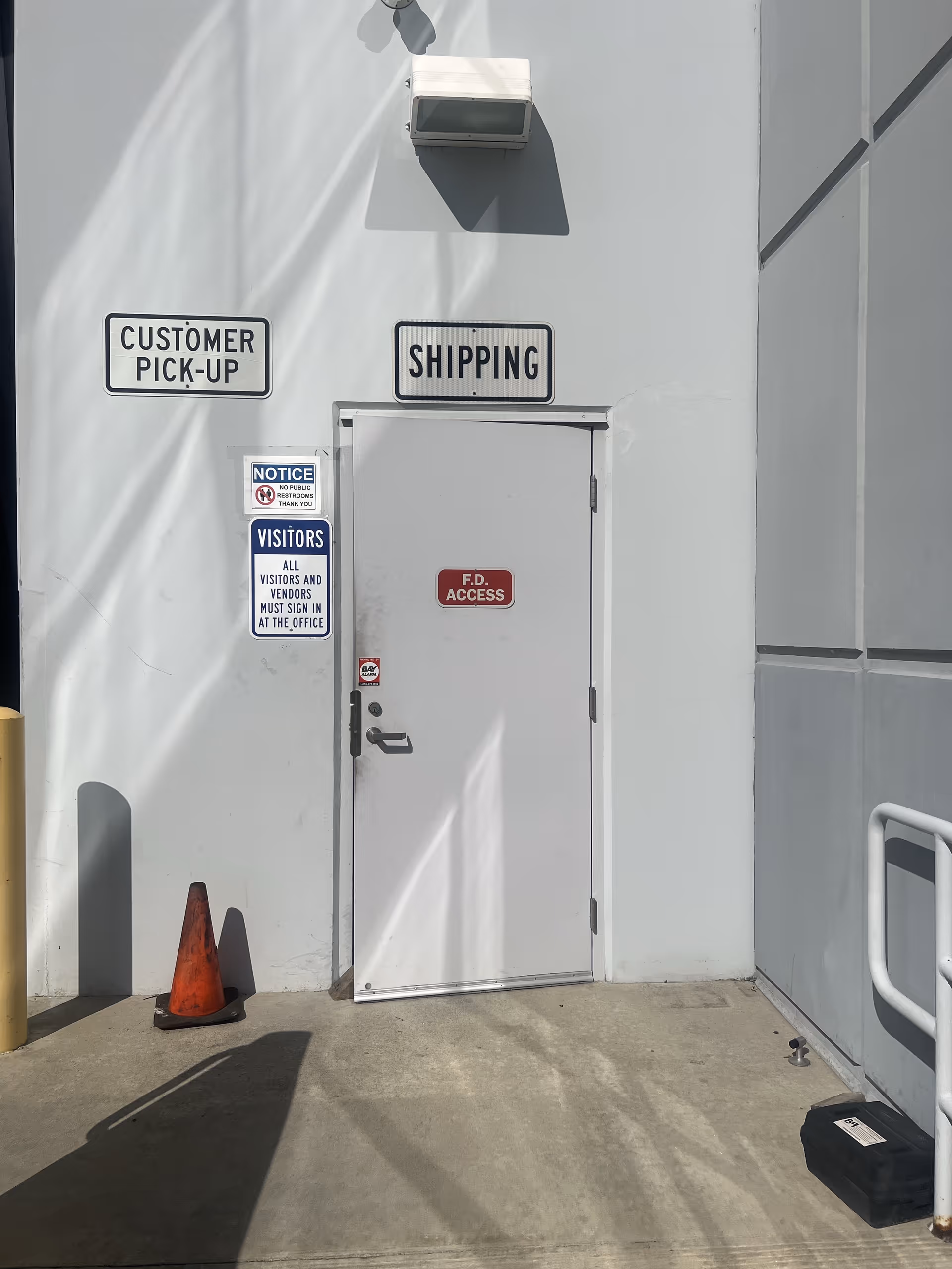 Exterior wall with a shipping door, customer pickup and visitors signs, and a cone.