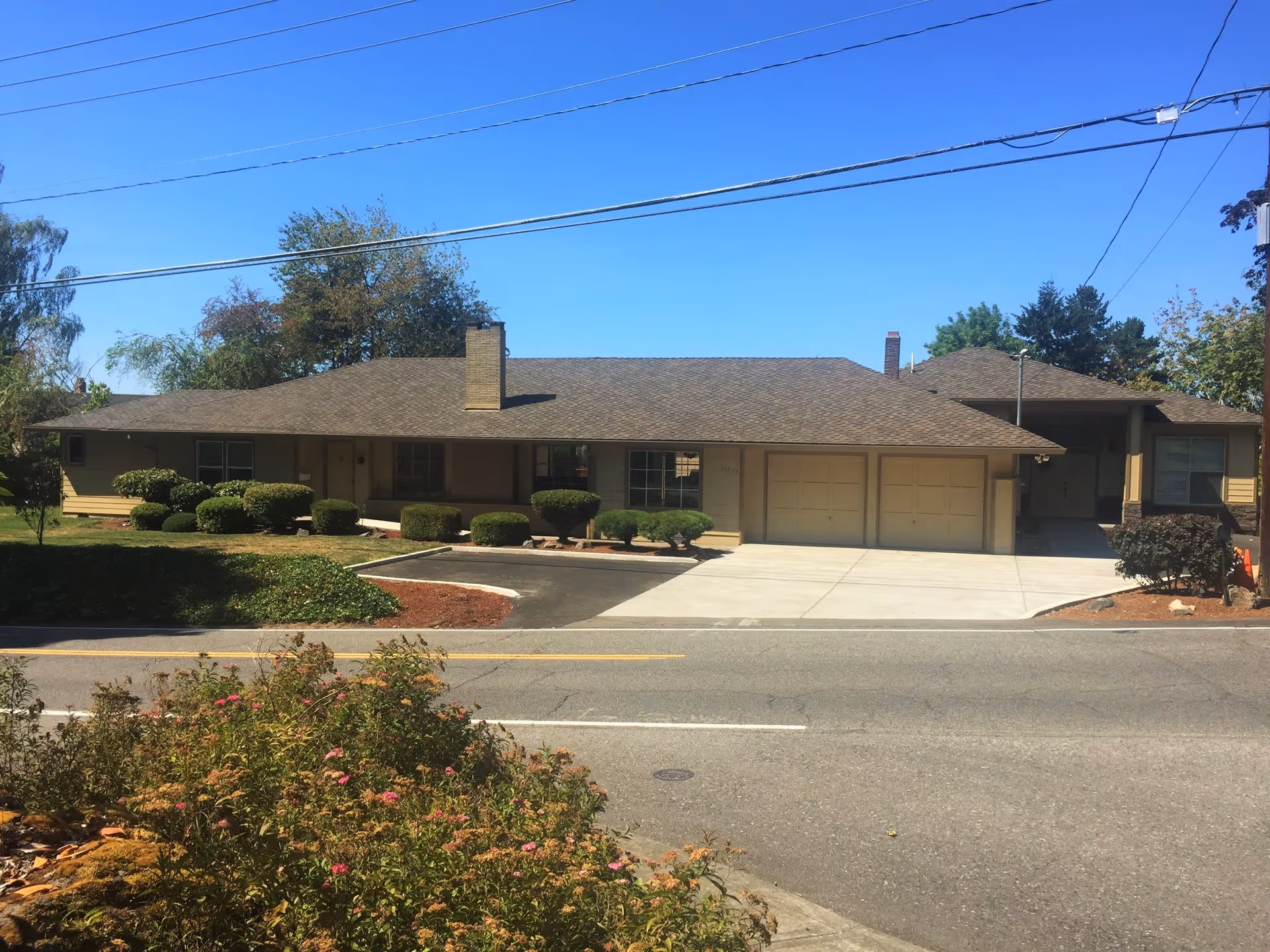 Single-story beige building with an attached two-car garage, trimmed hedges, driveway and a street in the foreground under a clear blue sky.