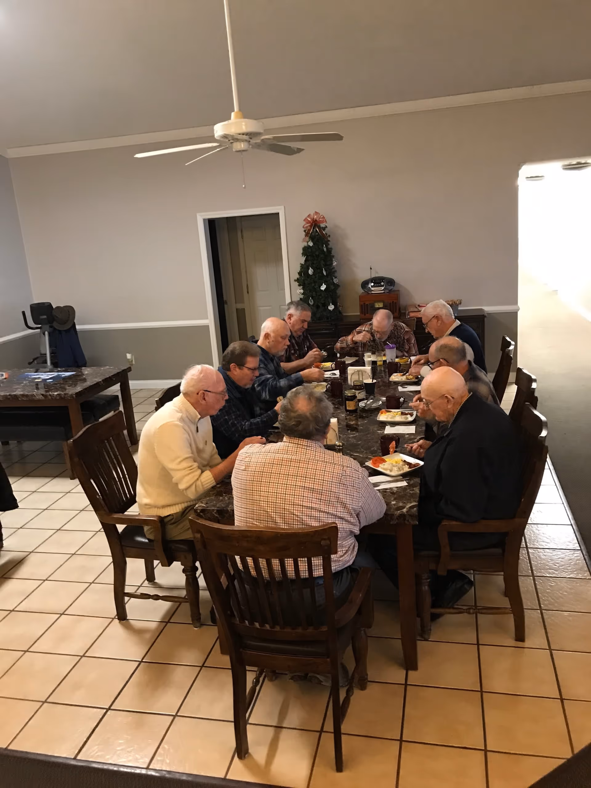 A group of elderly men sitting around a large dining table eating a meal in a communal dining room.