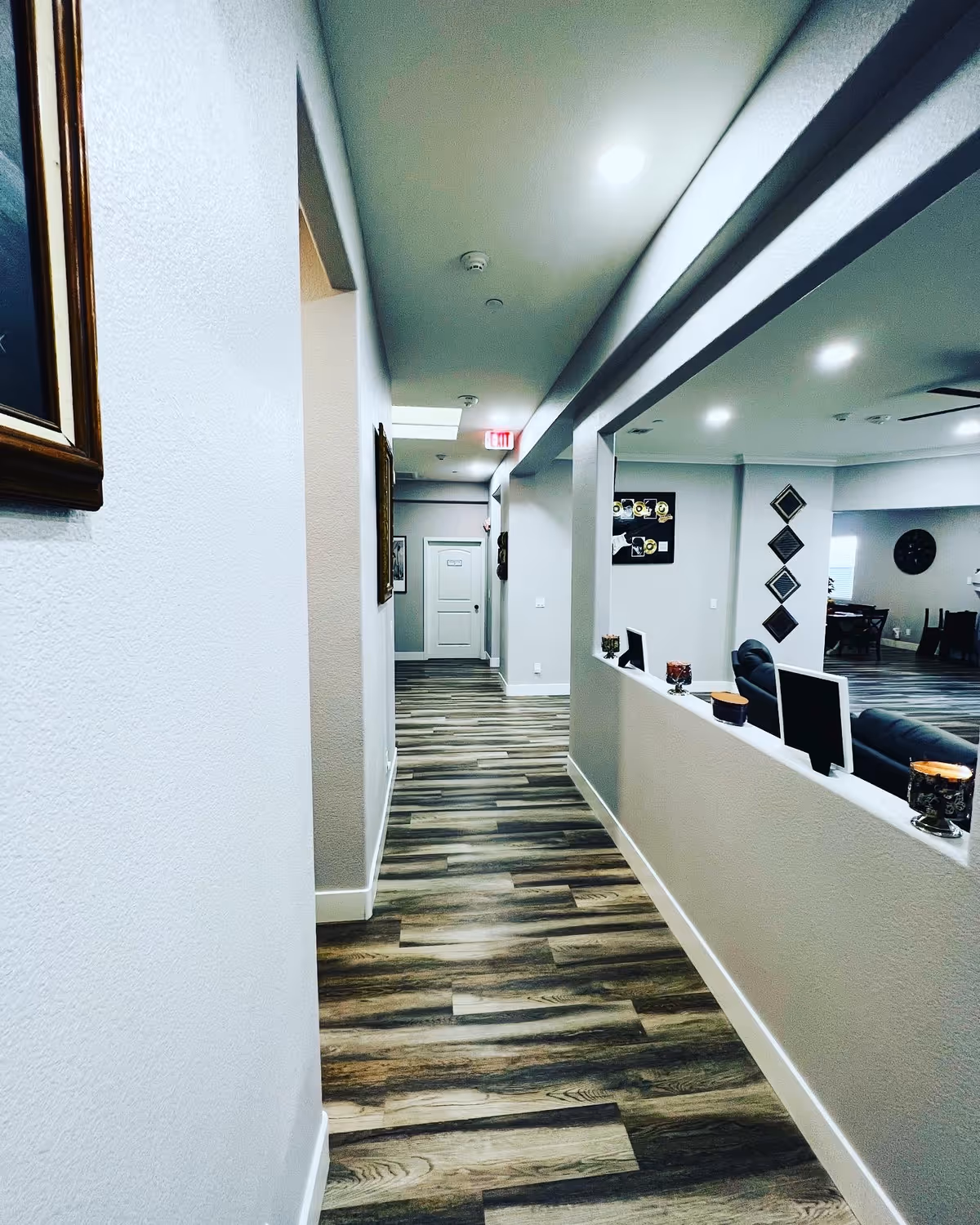 Interior hallway of EZ Years Retirement Living with wood-patterned flooring, light gray walls, and recessed ceiling lights. The hallway leads to a white door at the end, and there is an open partition on the right side showing a seating area with chairs and wall decorations.