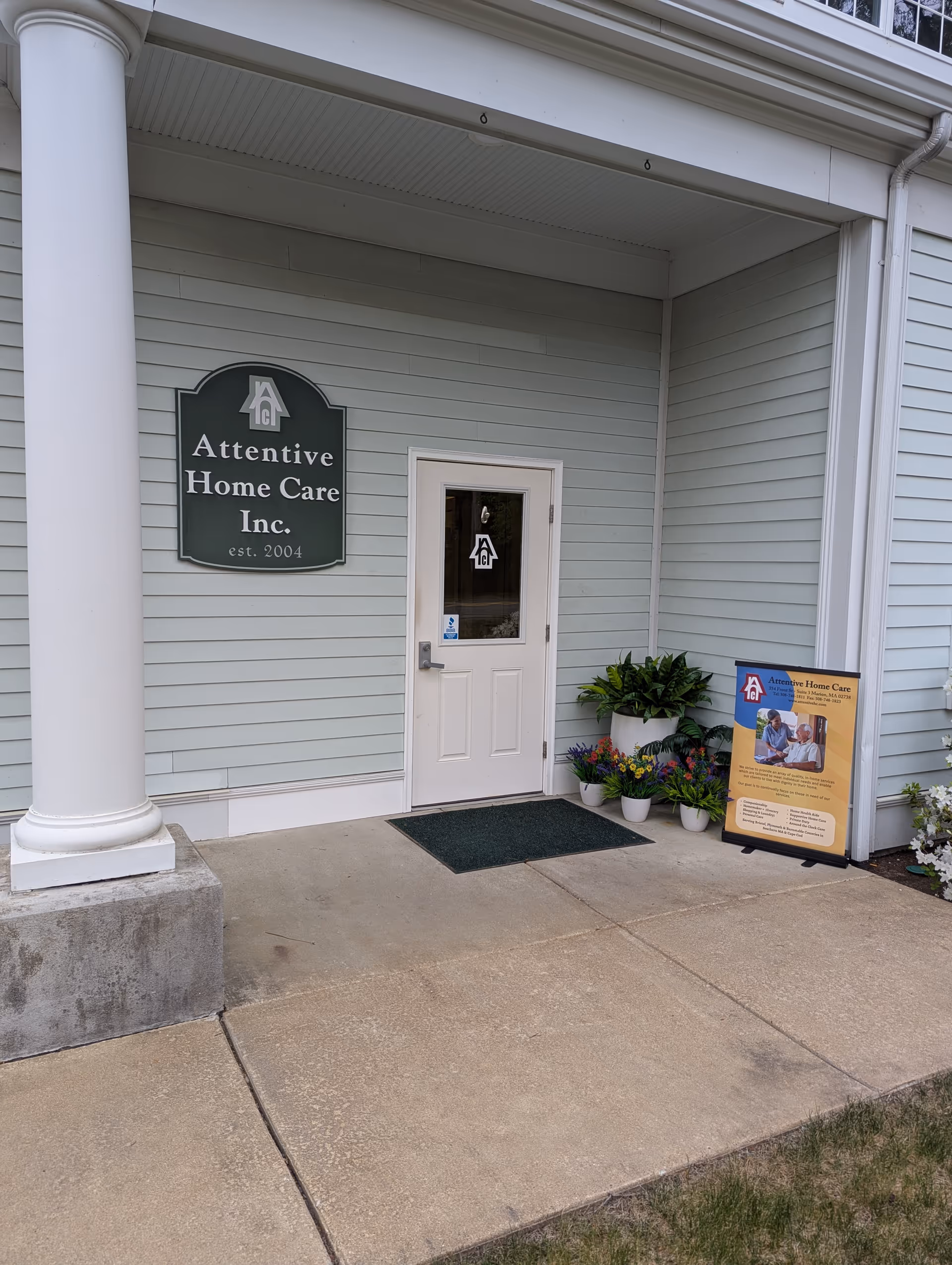 Front entrance of Attentive Home Care Inc. building with sign, door, and potted plants.
