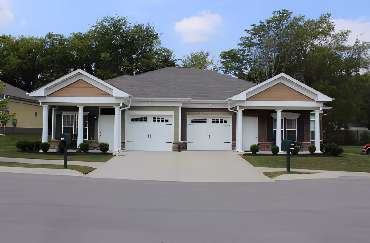 Single-story duplex-style senior living building with two garages, covered front porches, and a shared driveway surrounded by lawns and trees.
