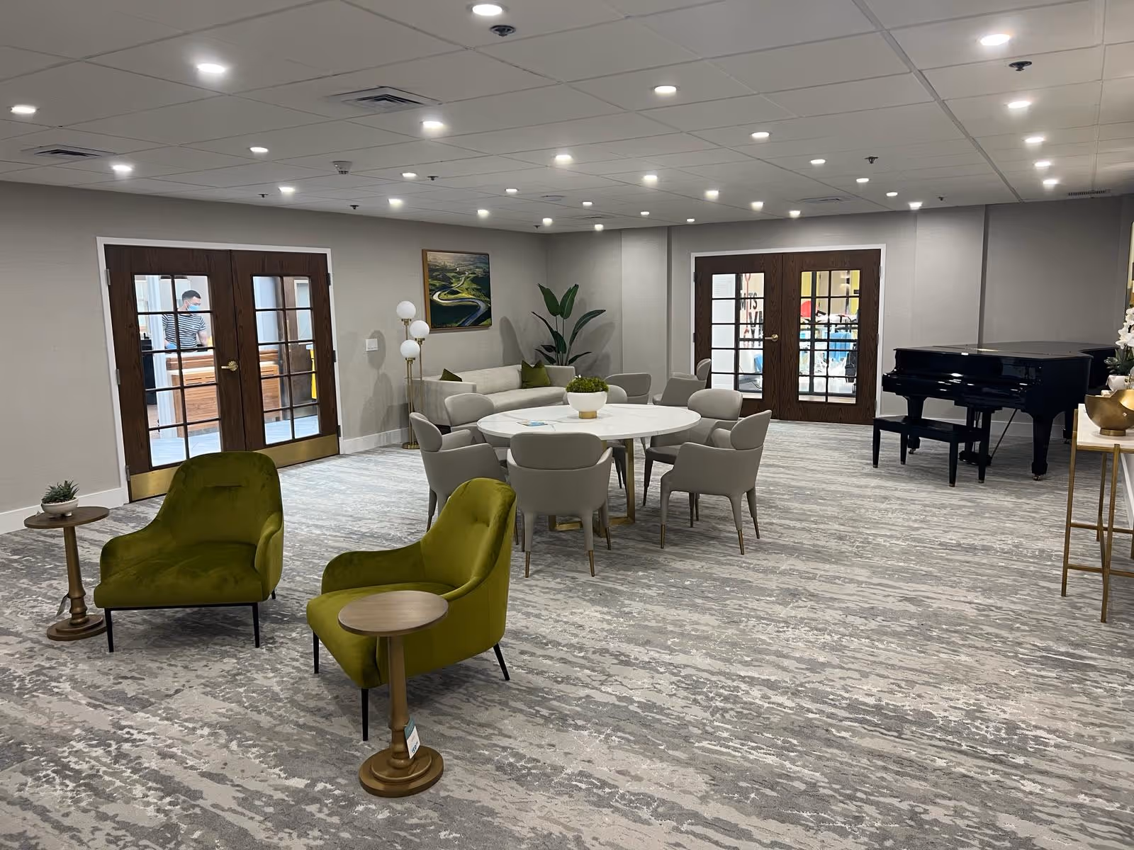 Spacious senior living common room with green lounge chairs, a round table with chairs, and a black grand piano near double French doors.