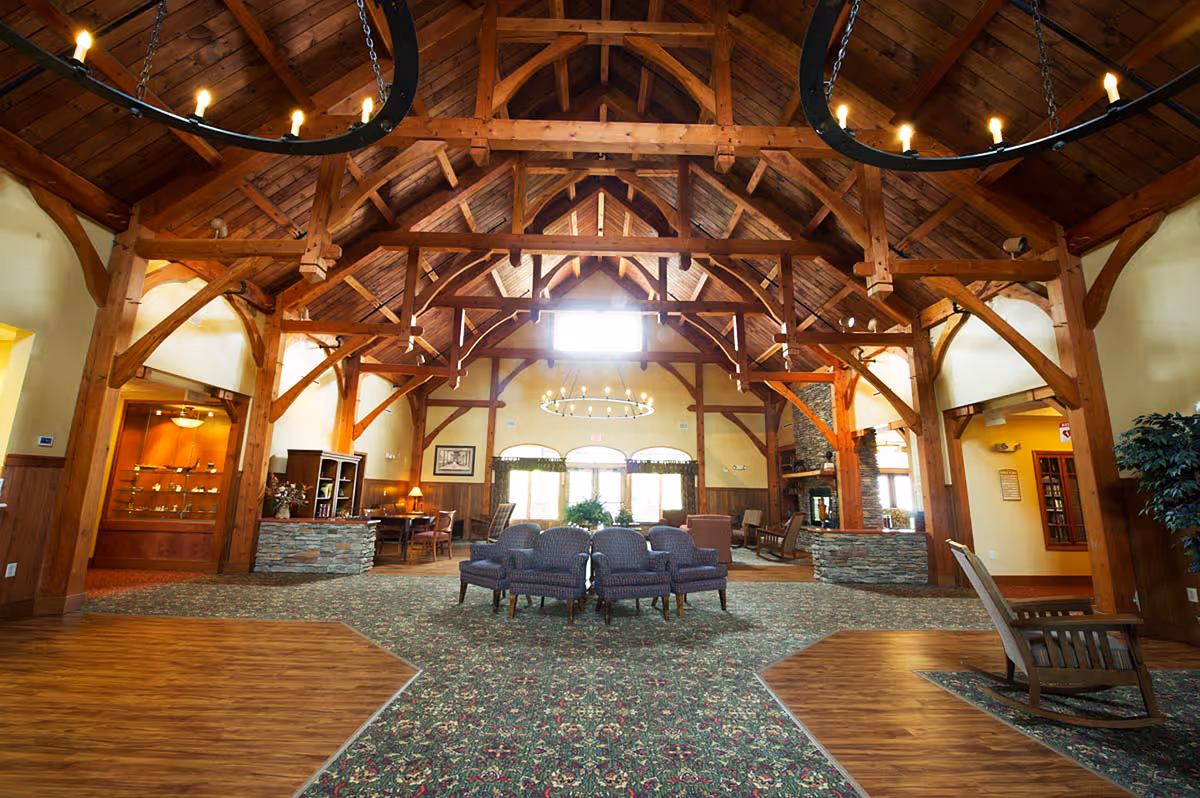 Spacious common area in a retirement community with high wooden vaulted ceilings, exposed beams, and large circular chandeliers. The room features a mix of carpet and wood flooring, several upholstered chairs arranged in the center, wooden furniture, and a stone fireplace on the right side. Large windows at the far end allow natural light to fill the space.