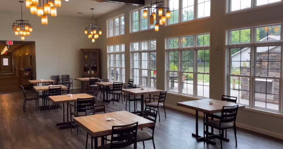 Bright dining room at Canterbury Arms Assisted Living with wooden tables, chairs, and large windows.