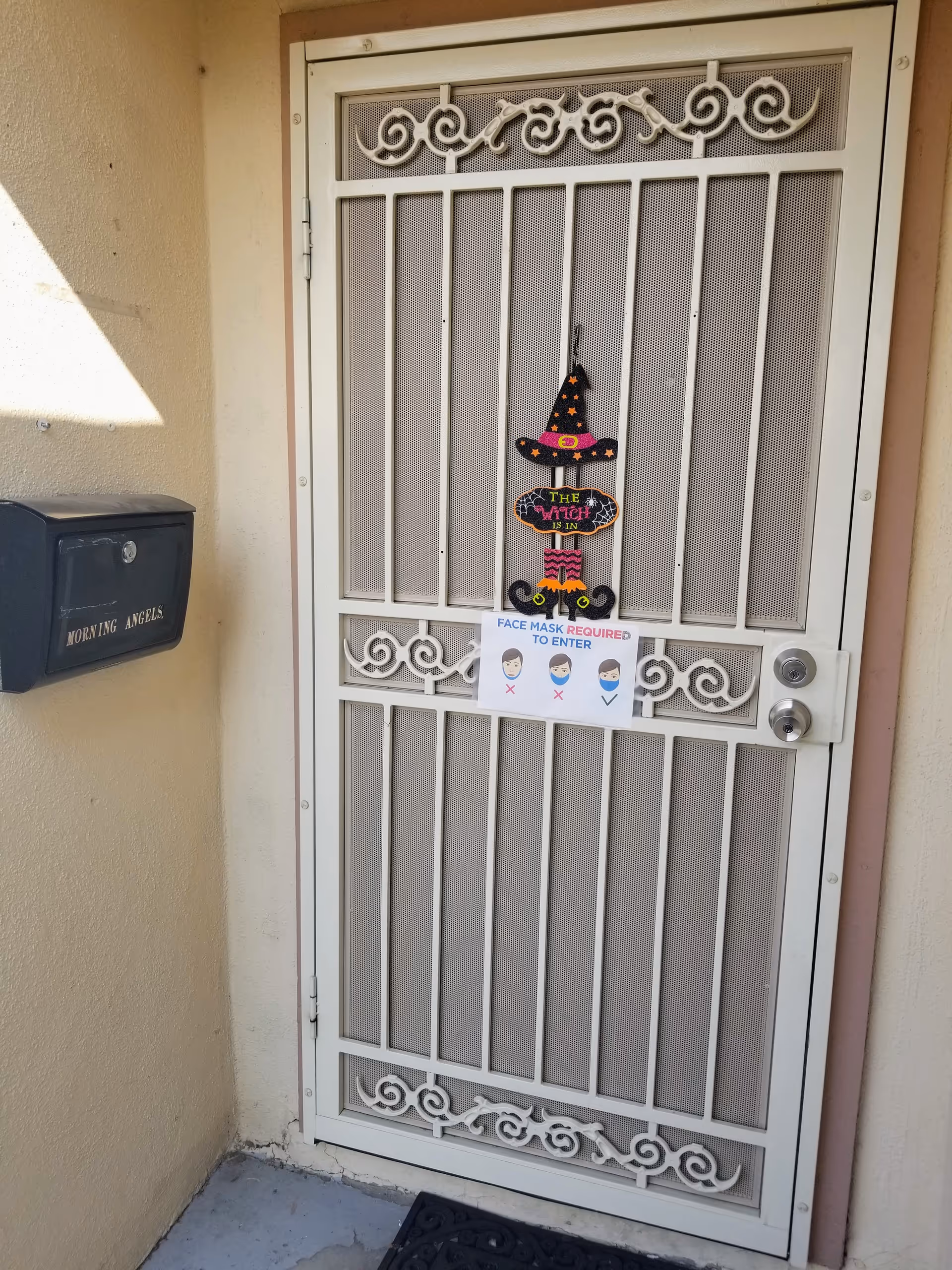 Exterior metal security door with decorative scrollwork, Halloween witch sign, and Morning Angels mailbox.