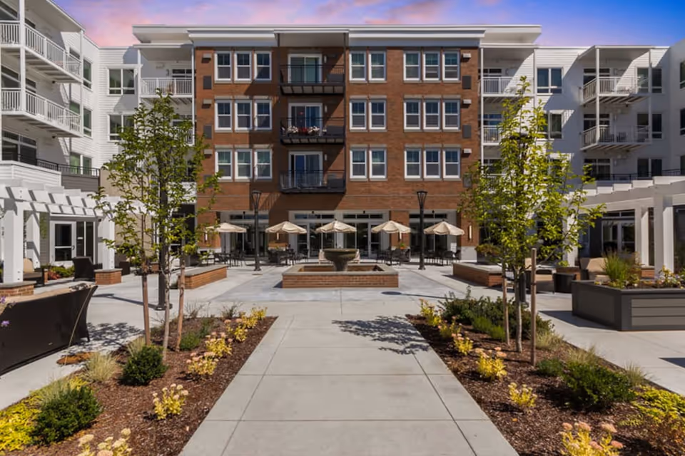 Outdoor central courtyard with a paved walkway, planted beds, seating areas with umbrellas, and a multi-story brick residential building with balconies in the background.