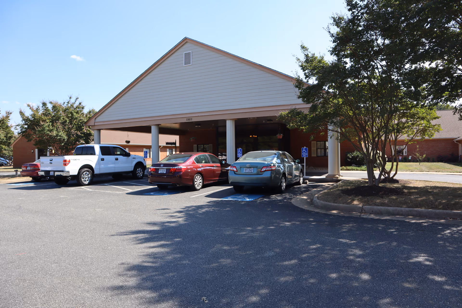Front exterior view of a single-story brick building with a covered entrance supported by white columns. Several cars are parked in front, including two in handicapped parking spaces. Trees and a clear blue sky are visible around the building.