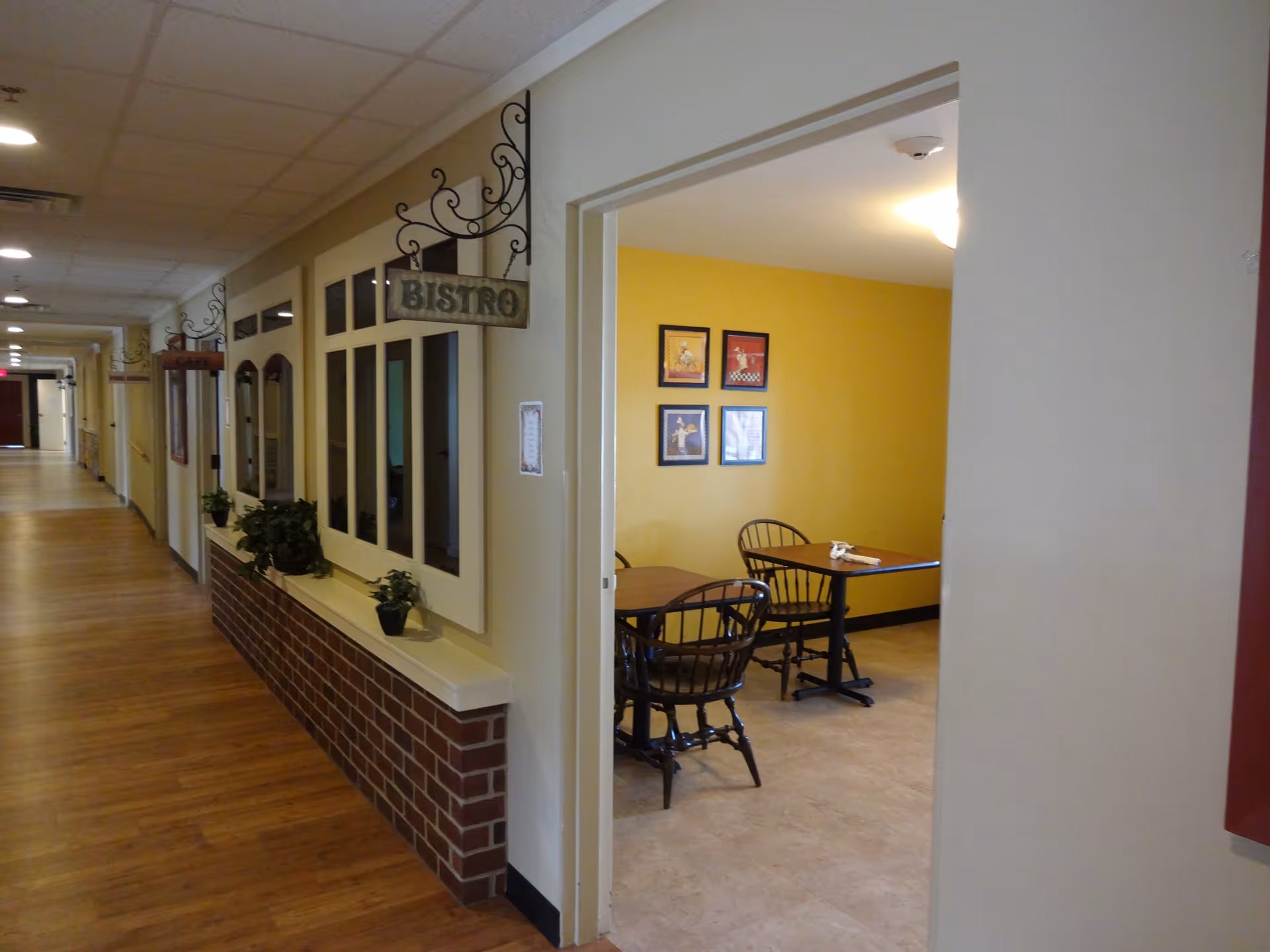 Interior view of a hallway in a senior living facility with wooden flooring and a wall decorated with small plants and windows. A sign hanging from the wall reads 'BISTRO'. To the right, there is an open doorway leading to a small dining area with yellow walls, a table, and wooden chairs. Four framed pictures are hung on the yellow wall.