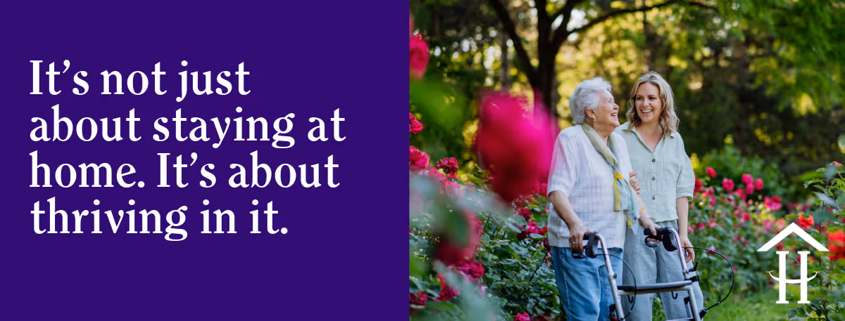 Elderly woman with walker walks beside a caregiver in a flower-filled outdoor garden.