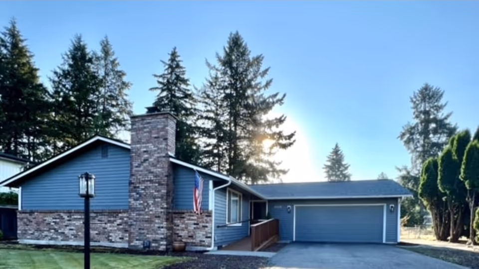 Blue single-story facility with brick chimney, driveway, two-car garage, flag, and trees.