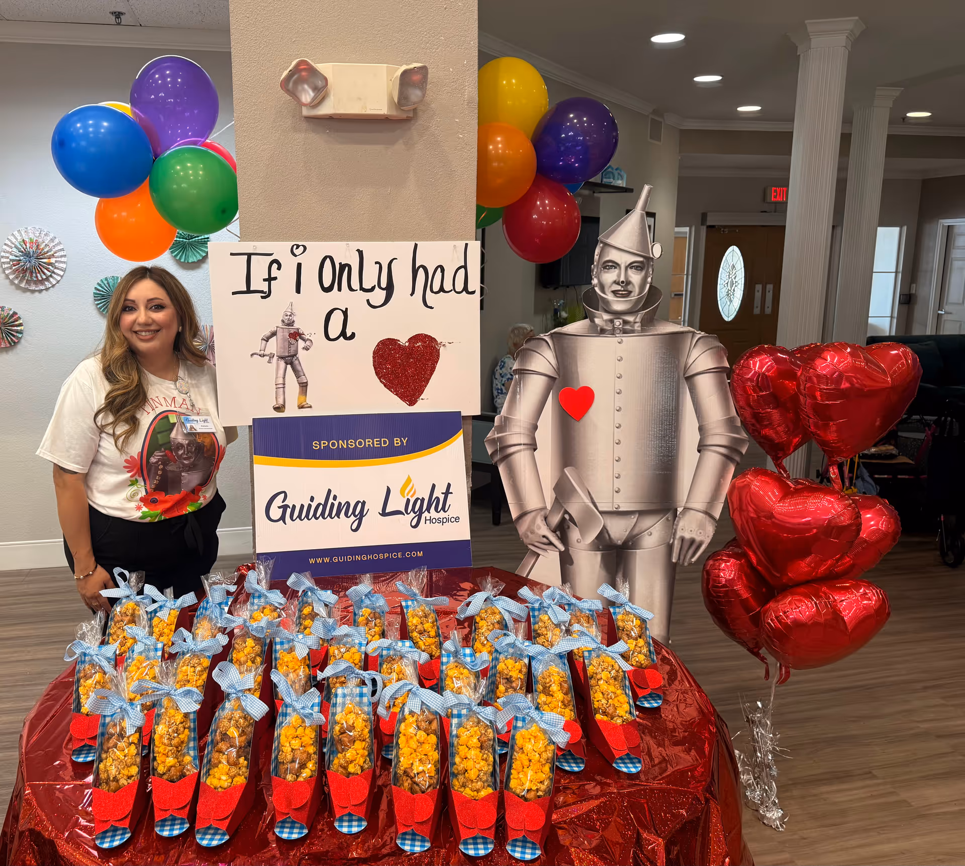 Smiling woman stands beside a tin man cutout, balloons, and gift bags at an indoor event.