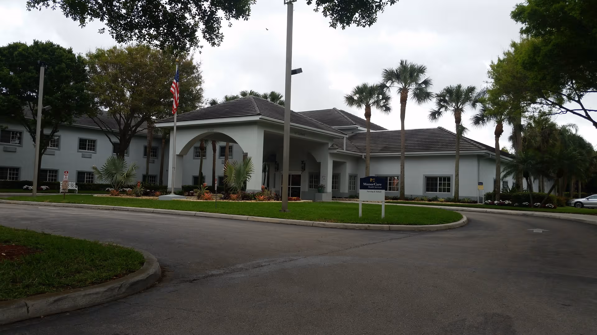White single-story senior facility with porte-cochère, palm trees, circular driveway, and flag outside.