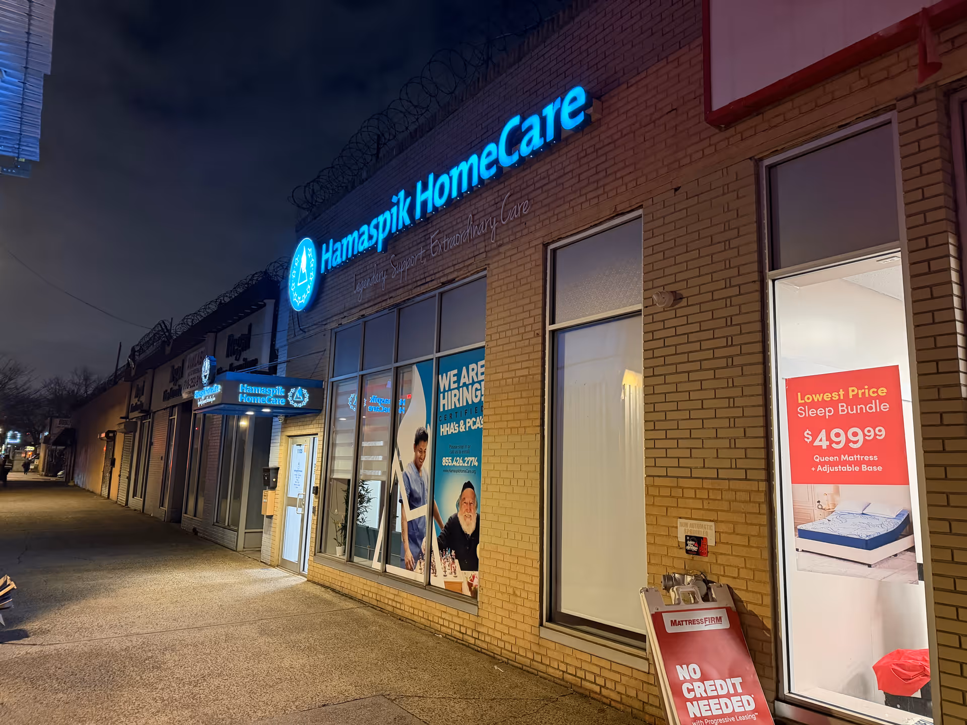 Night exterior of Hamaspik HomeCare storefront featuring blue illuminated sign along brick facade.